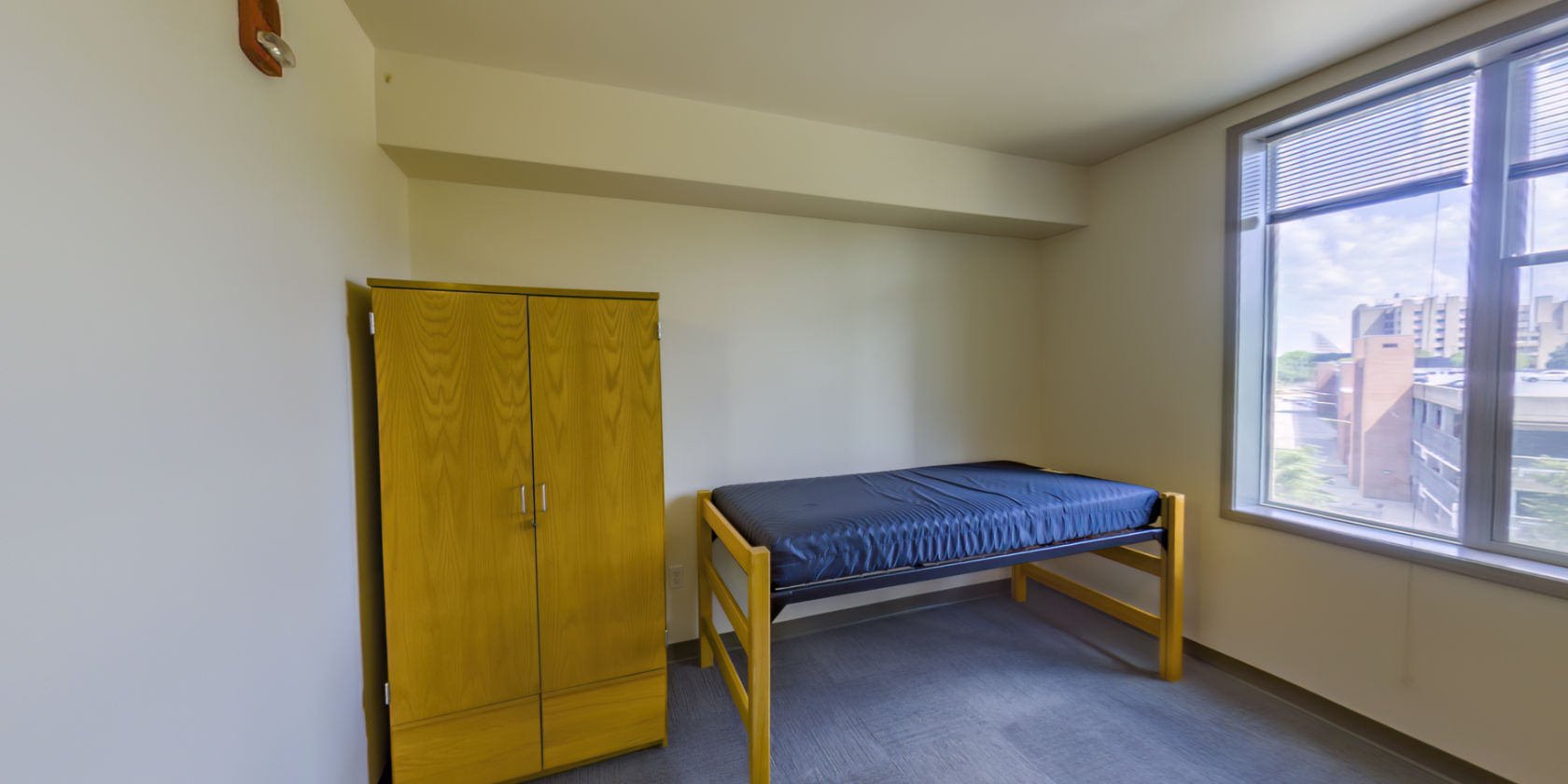 This single bedroom has a carpeted floor, a single bed with space below for storage, and closet storage with a small latch for a padlock. There are shaded windows with a view overlooking the campus.