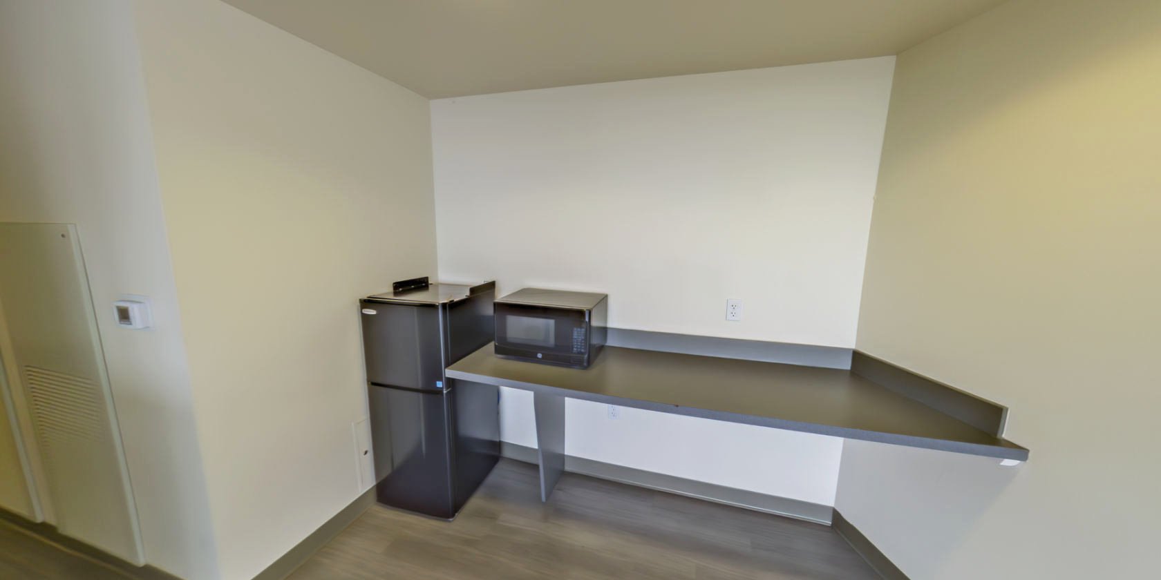 This common area has laminate flooring and an open countertop space with a microwave. Next to the countertop is a small refrigerator.