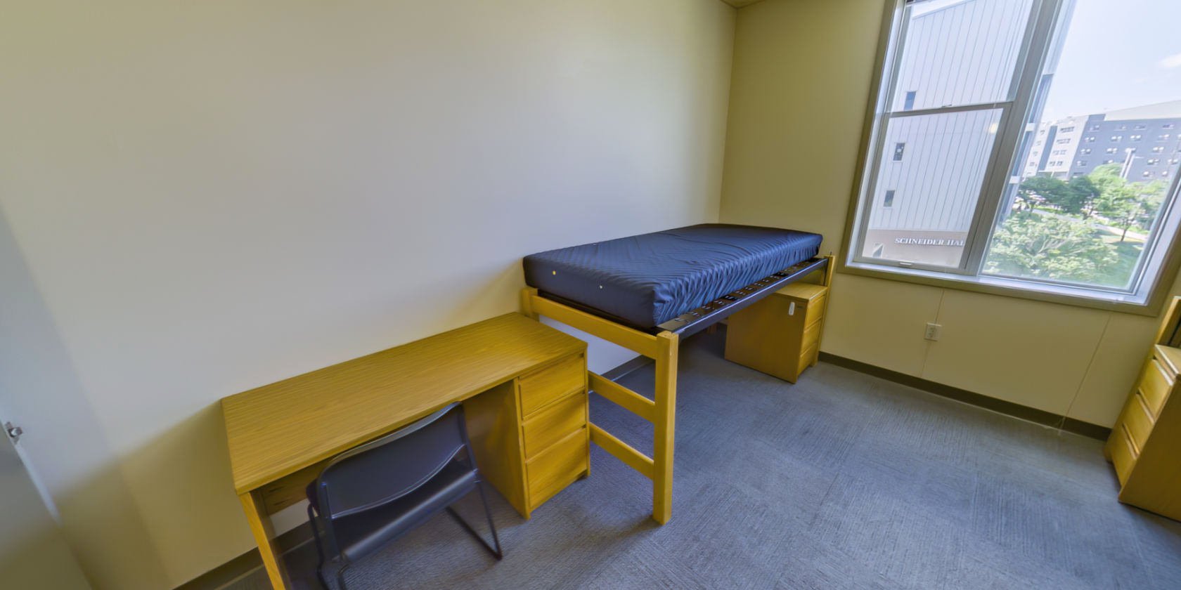 This first double bedroom has a carpeted floor, a single bed with space below for bedside storage, and a desk with drawers. There are shaded windows with a view overlooking the campus.