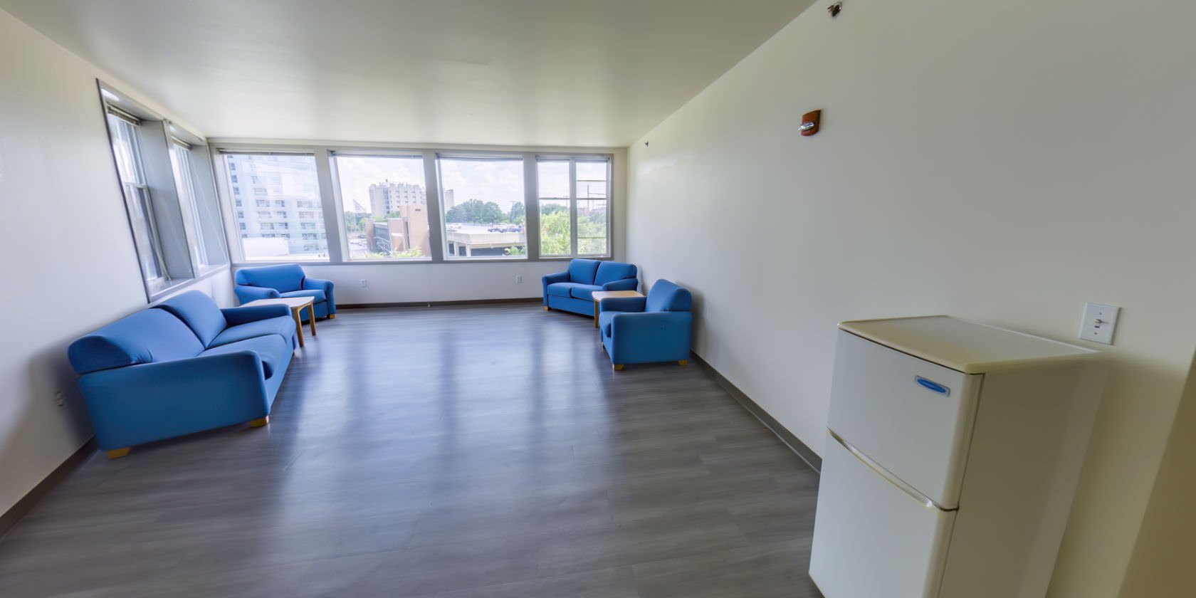 This side of the common area has two sofas, two chairs, and two small side tables. There are shaded windows with a panoramic view of the campus. Near the entrance to the hallway is a second small refrigerator.