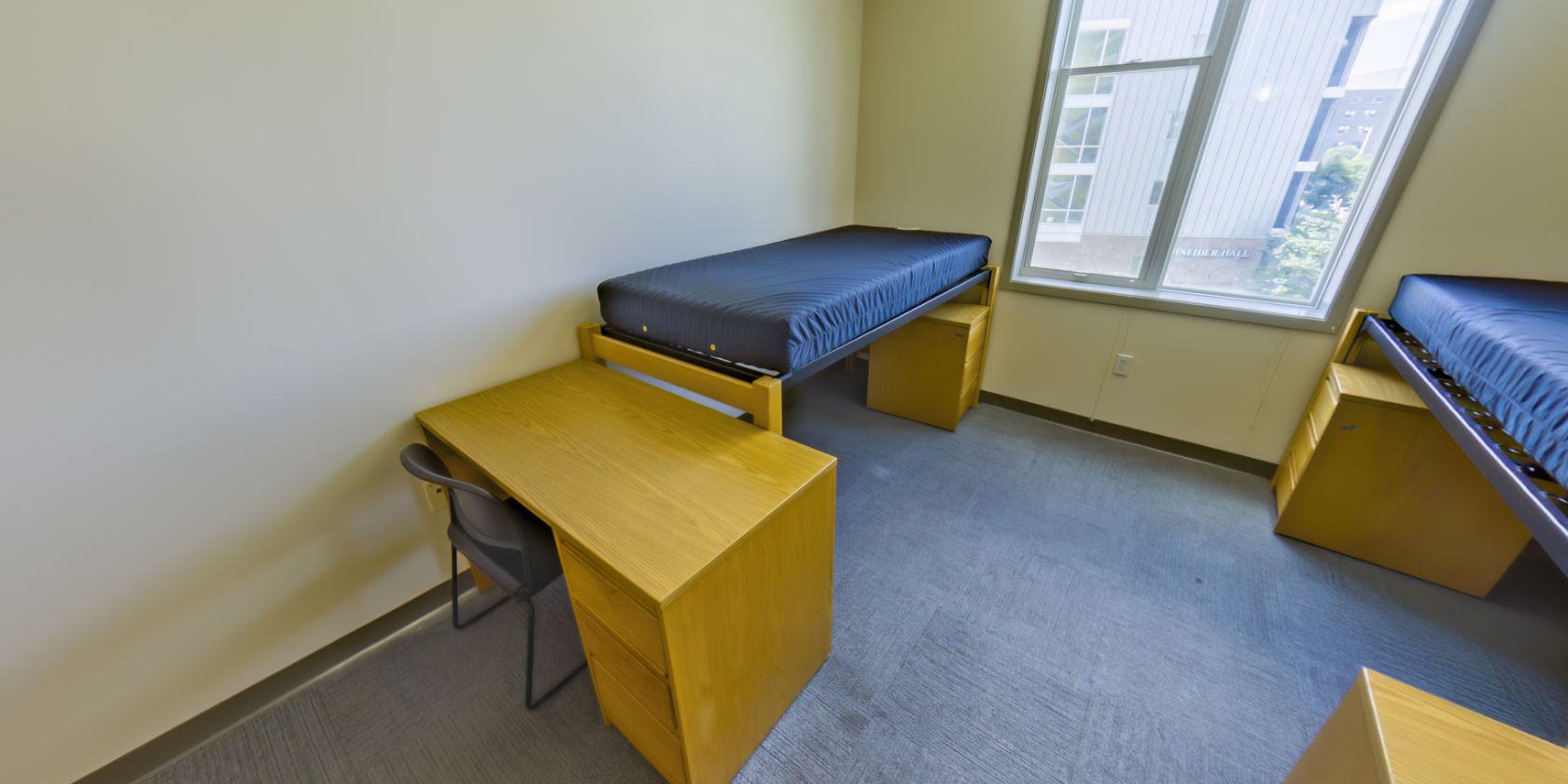 This second double room has a carpeted floor, a single bed with space below for bedside storage, and a desk with drawers. There are shaded windows with a view overlooking the campus.