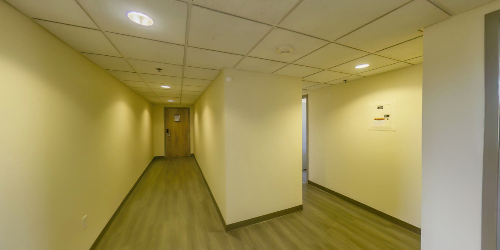 This virtual tour begins in a hallway inside the entrance to a suite. The hallway has laminate flooring and connects to a second hallway.