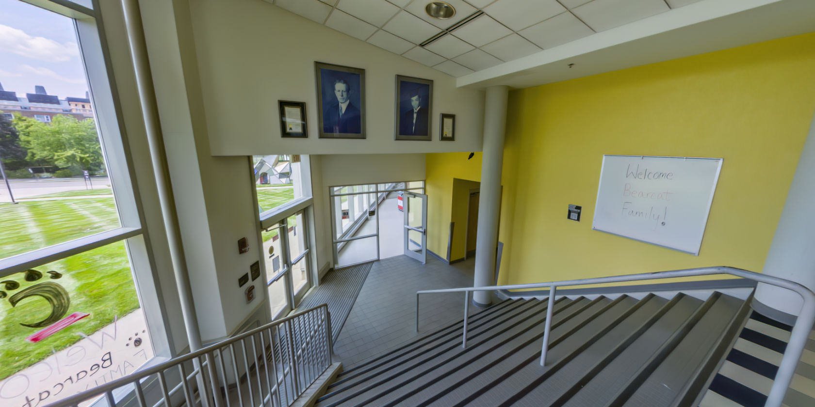 This side of the lobby has a set of non-slip stairs with handrails leading down to more spaces. There are two portraits hanging above the space and a whiteboard on the wall. There are windows with a panoramic view of the campus.