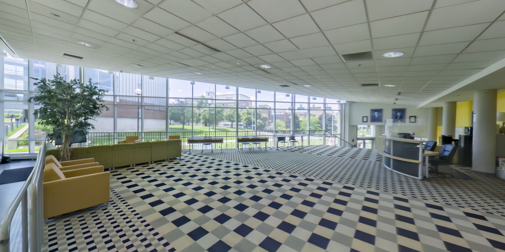 This virtual tour begins inside Turner Hall Lobby, in an elevated space with a carpeted floor and a reception desk. Off to one side is a sitting area with a sofa, several chairs, and a TV. Next to the sitting area, there are three long tables with chairs. There is a non-slip accessible ramp with a handrail leading down to an exit. There are windows outlining the space with a panoramic view of the campus.
