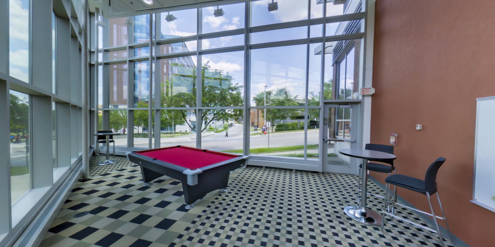 This side of the breakout room has a lounge area with a carpeted floor, three high-top tables with seating, and a whiteboard on the wall. There is a pool table in the middle and windows outlining the space with a panoramic view of the campus.