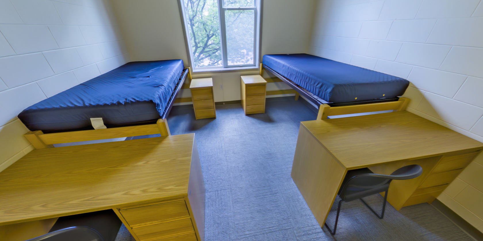 This first carpeted bedroom has two single beds with bedside storage, two desks with drawers, and shaded windows with a view overlooking the campus. There is space below the beds for additional storage.