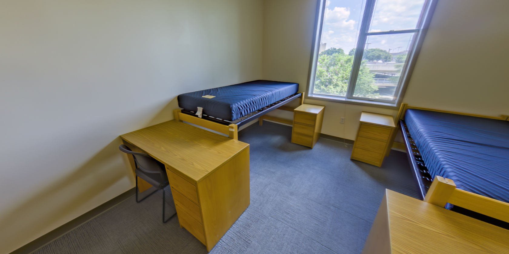 This second carpeted room has a single bed with space below for storage and a desk with drawers. There is bedside storage next to the bed and shaded windows with a view overlooking the campus.
