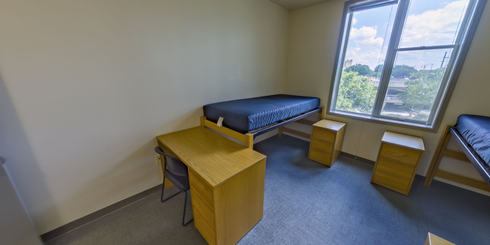 This first carpeted bedroom has a single bed with space below for storage and a desk with drawers. There is bedside storage next to the bed and shaded windows with a view overlooking the campus.