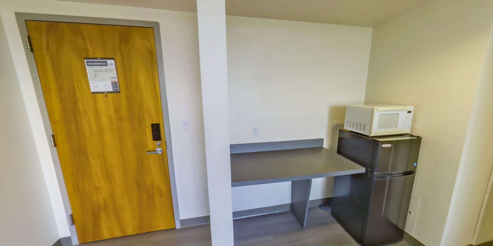 This virtual tour begins inside the entrance to a suite with laminate flooring. Near the entrance is a space containing a countertop and a small refrigerator with an attached microwave.