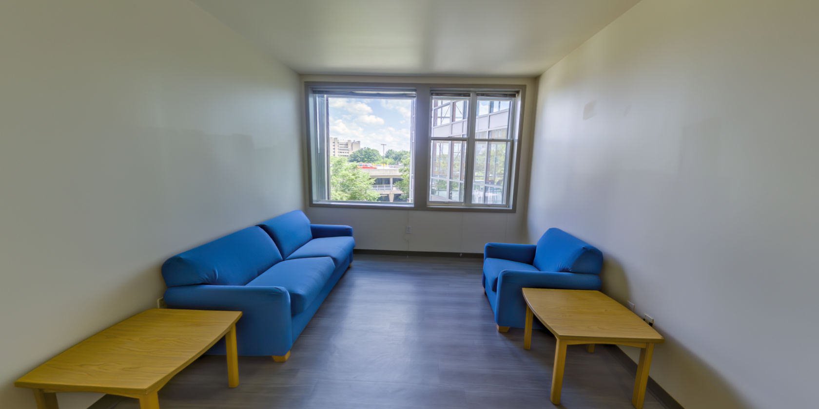 This side of the entrance connects to the common area with laminate flooring and a sofa with a side table. Across from the sofa is a chair and a second side table. There are shaded windows with a view overlooking the campus.